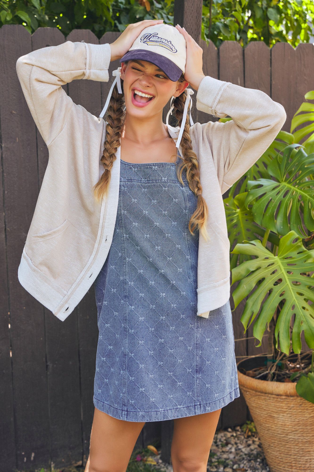 Woman wearing a denim dress, beige jacket, and cap against a wooden fence.