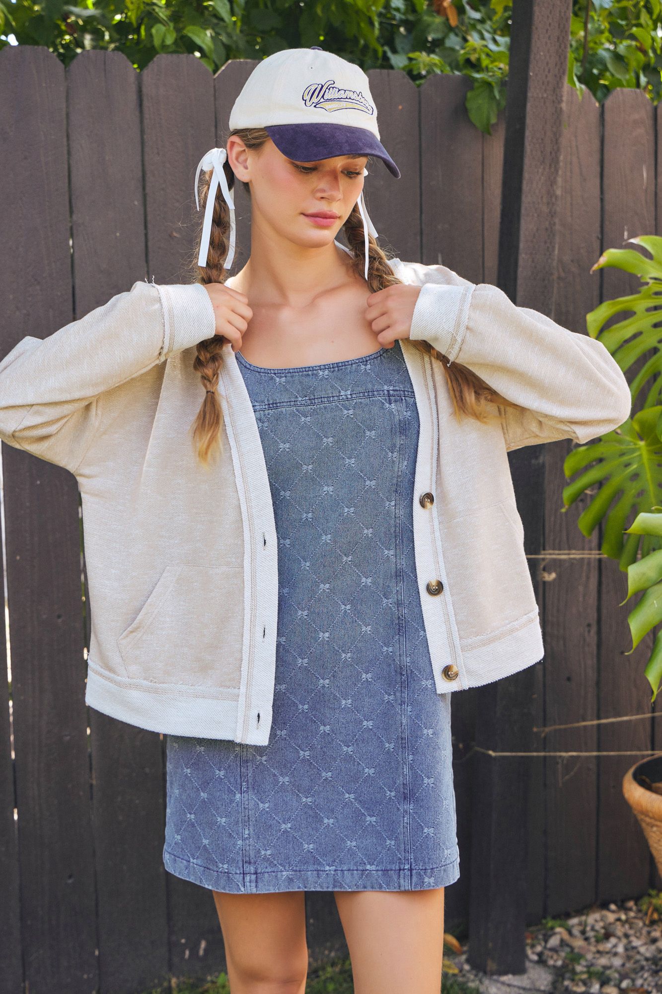 Woman wearing a denim dress, beige jacket, and cap against a wooden fence.