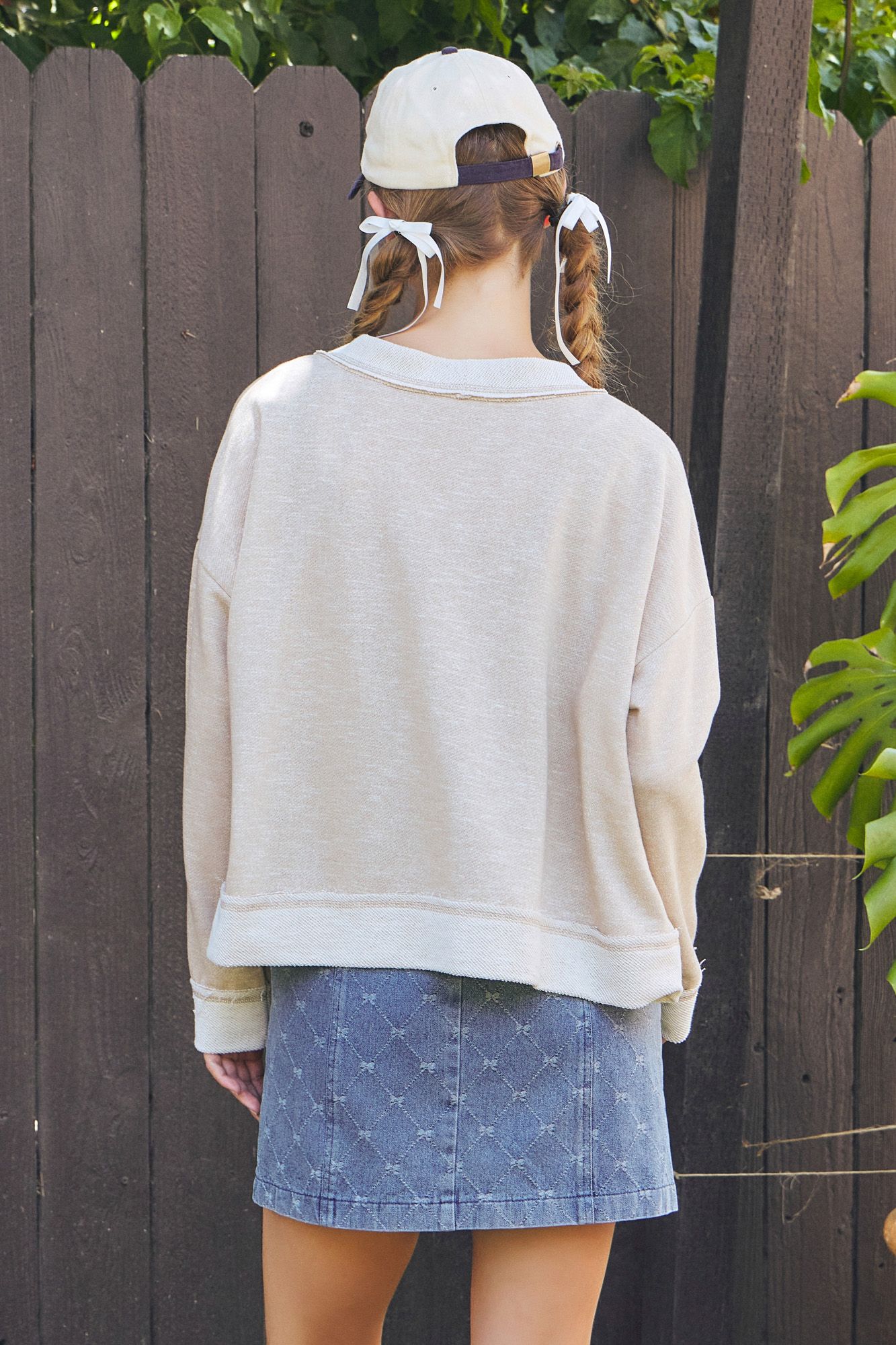Person wearing a beige sweater and blue denim skirt with a white cap, standing against a wooden fence.