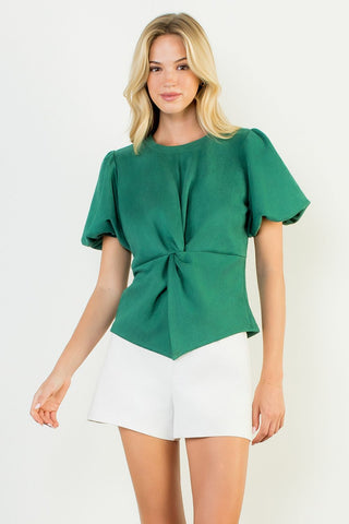 Woman wearing a green top with a knot detail and white shorts on a light background