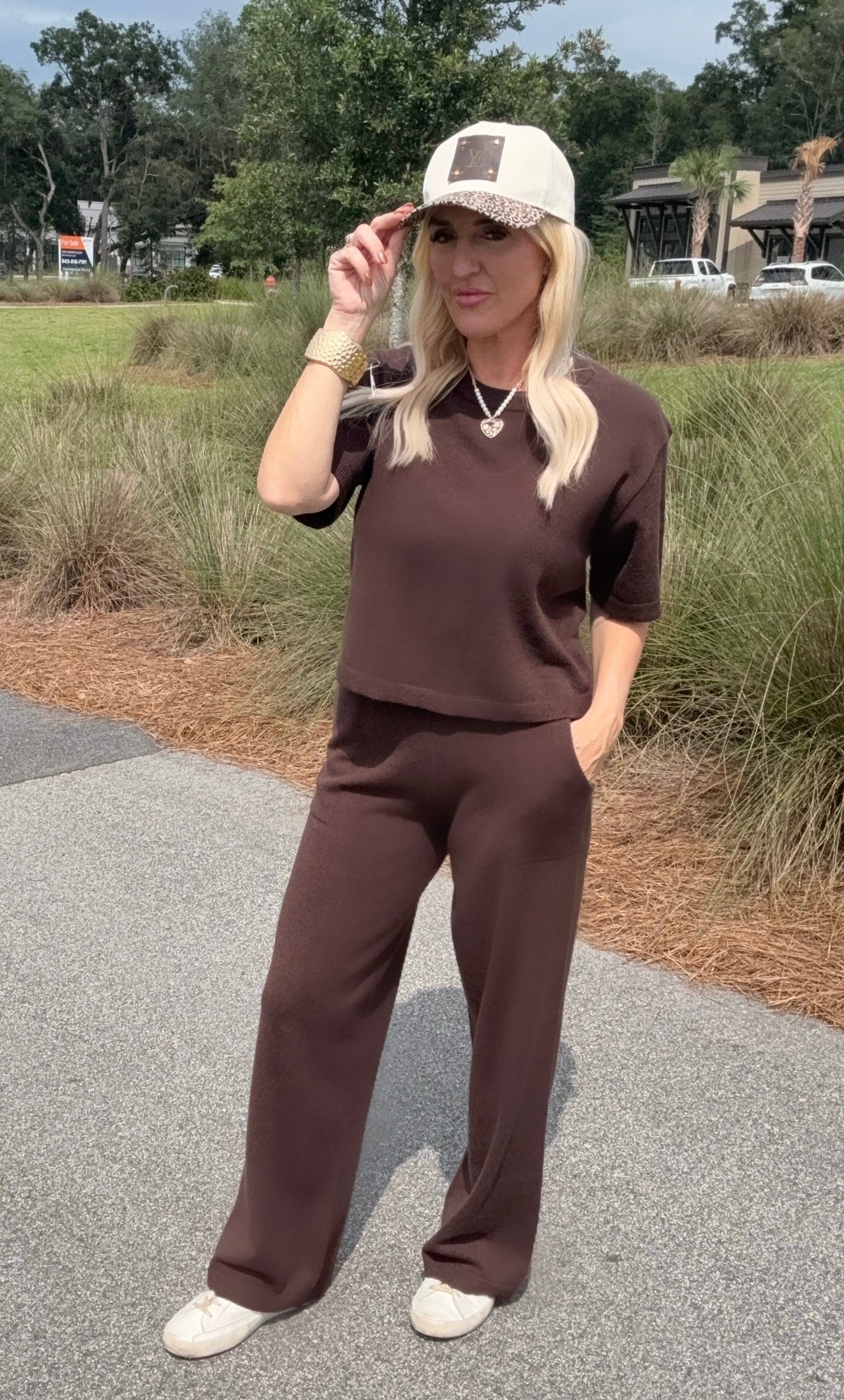 Woman in brown outfit and cap standing outdoors on a path with greenery.