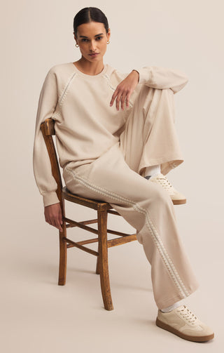 Woman sitting on a wooden chair wearing a beige outfit with a plain background