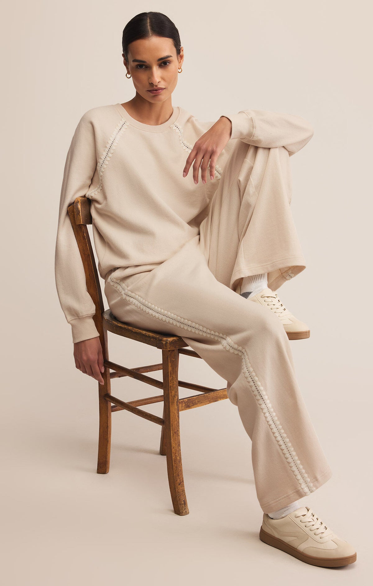 Woman sitting on a wooden chair wearing a beige outfit with a plain background