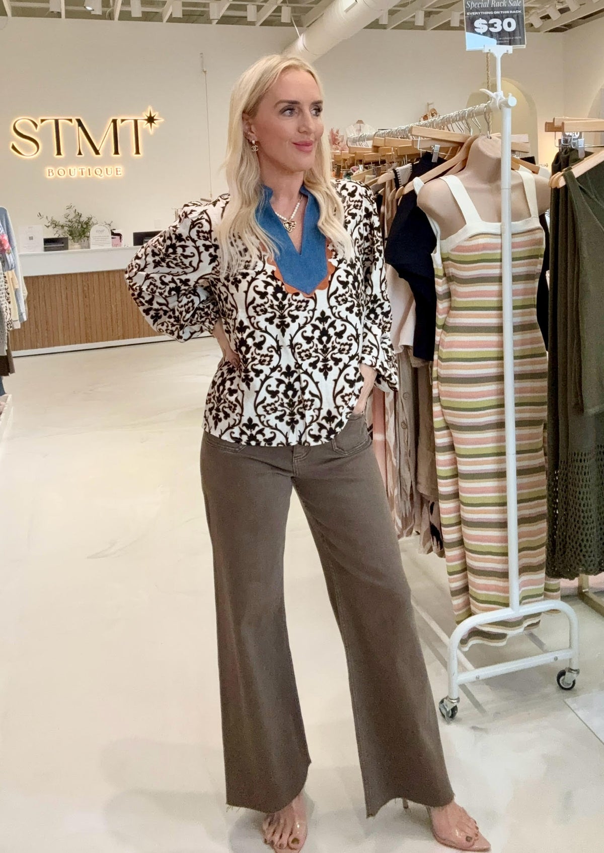 Woman in a patterned blouse and brown pants standing in a store with clothing racks and a 'STMT' sign.