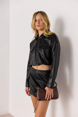 Woman wearing a black leather jacket and skirt holding a brown handbag against a white background