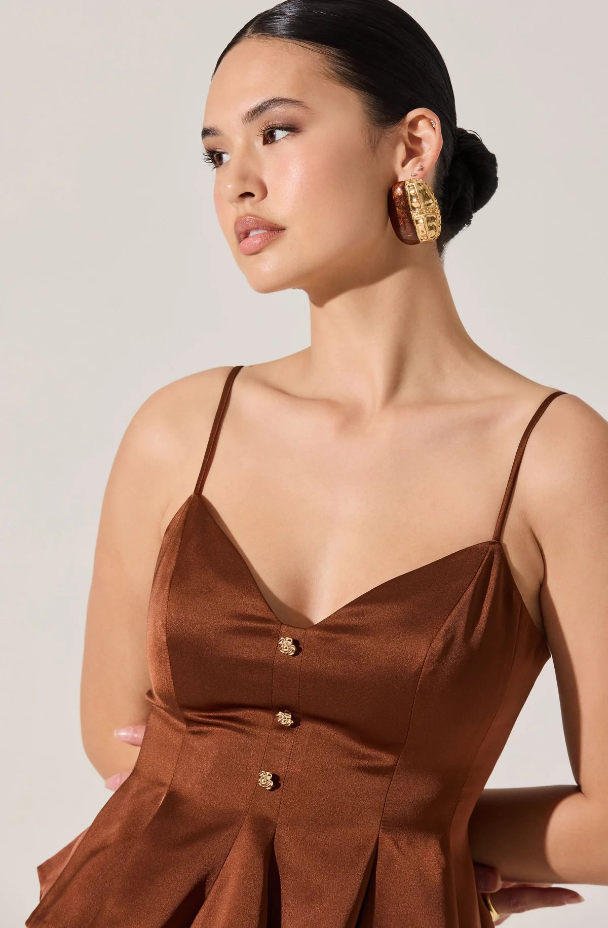 Woman wearing a brown dress with gold buttons and earrings against a plain background