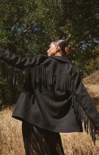 Person wearing a black fringed jacket in a natural setting with trees and dry grass.