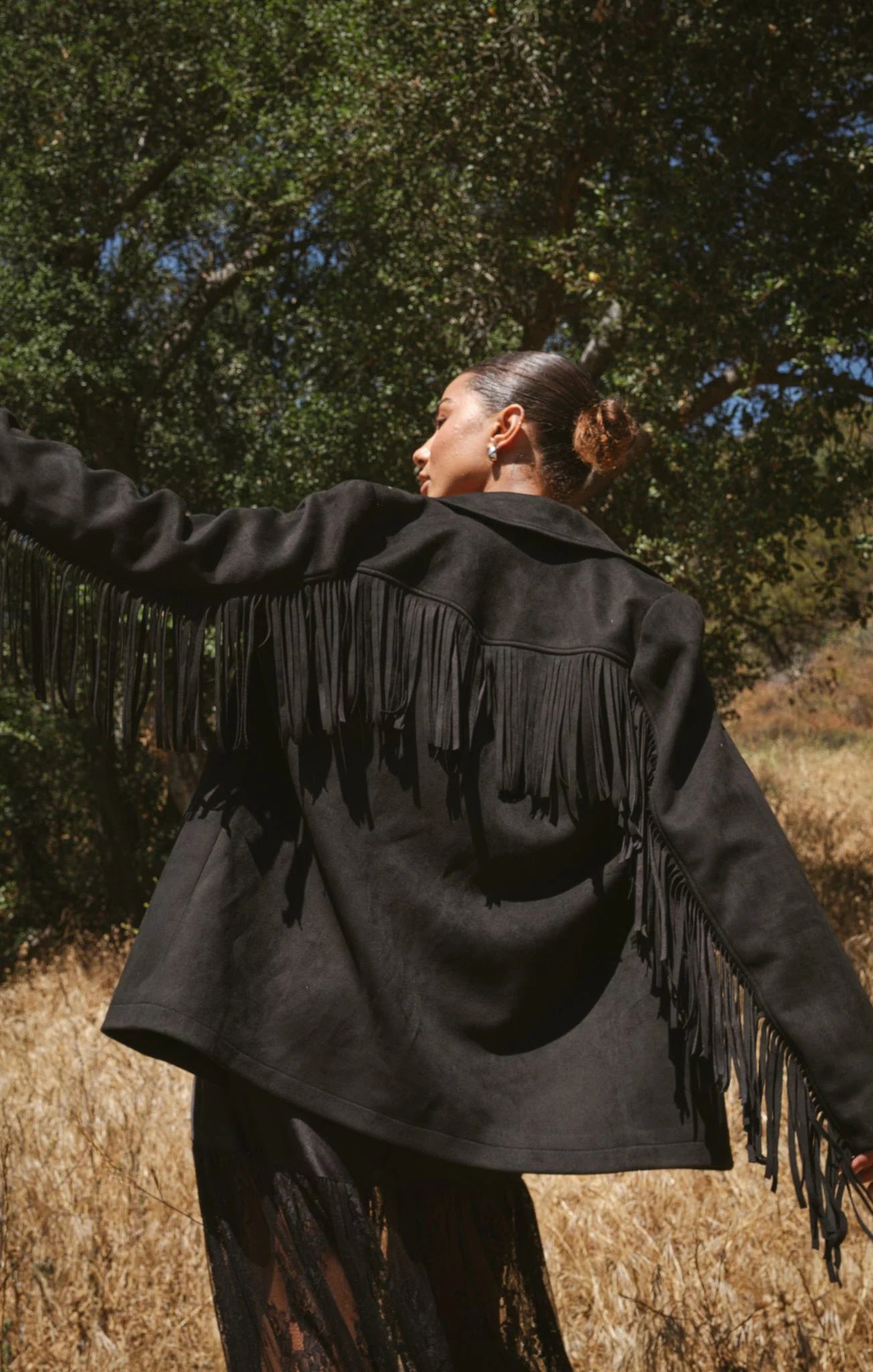 Person wearing a black fringed jacket in a natural setting with trees and dry grass.