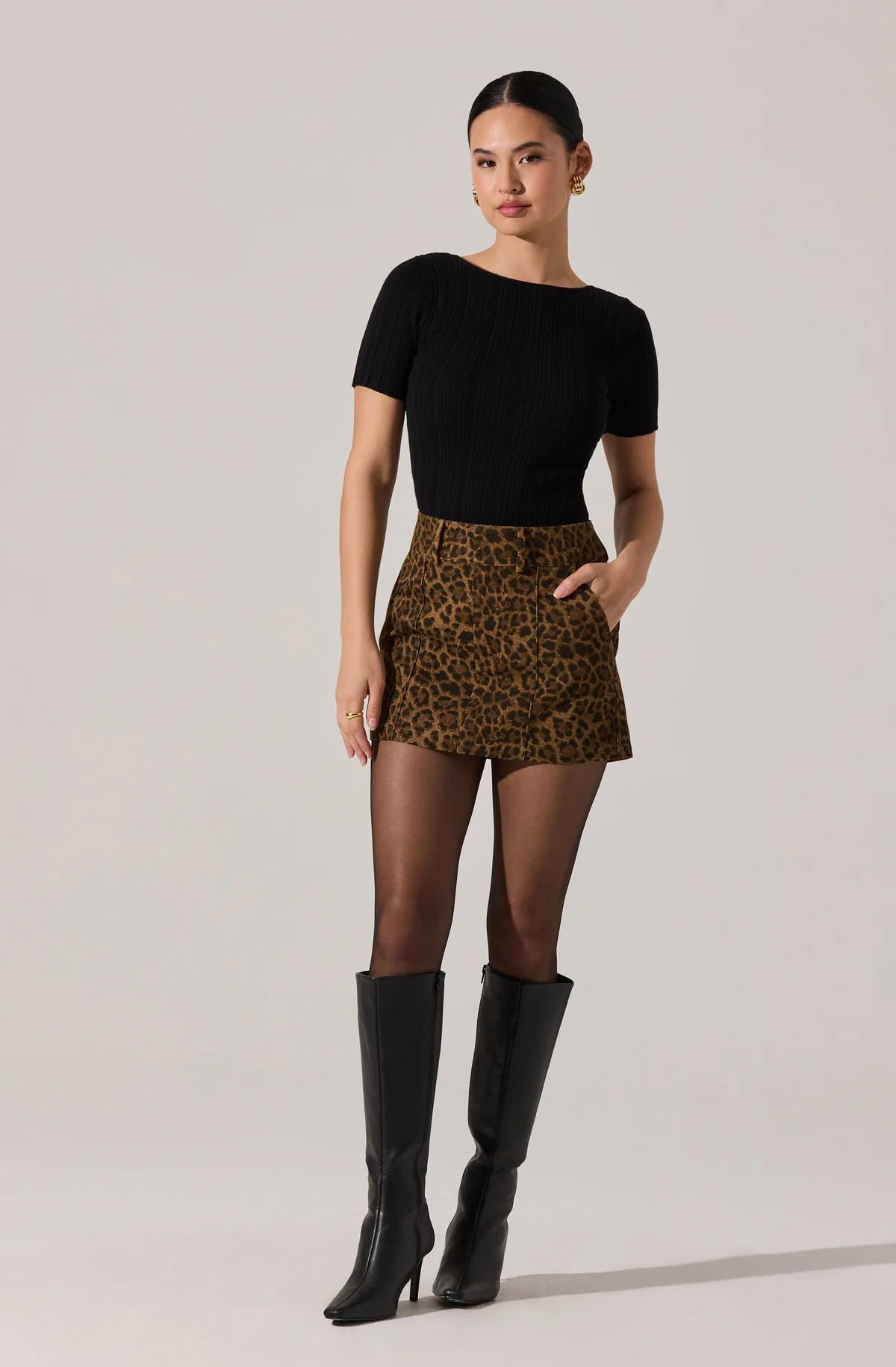 Woman wearing a black top, leopard print skirt, and black knee-high boots on a light gray background