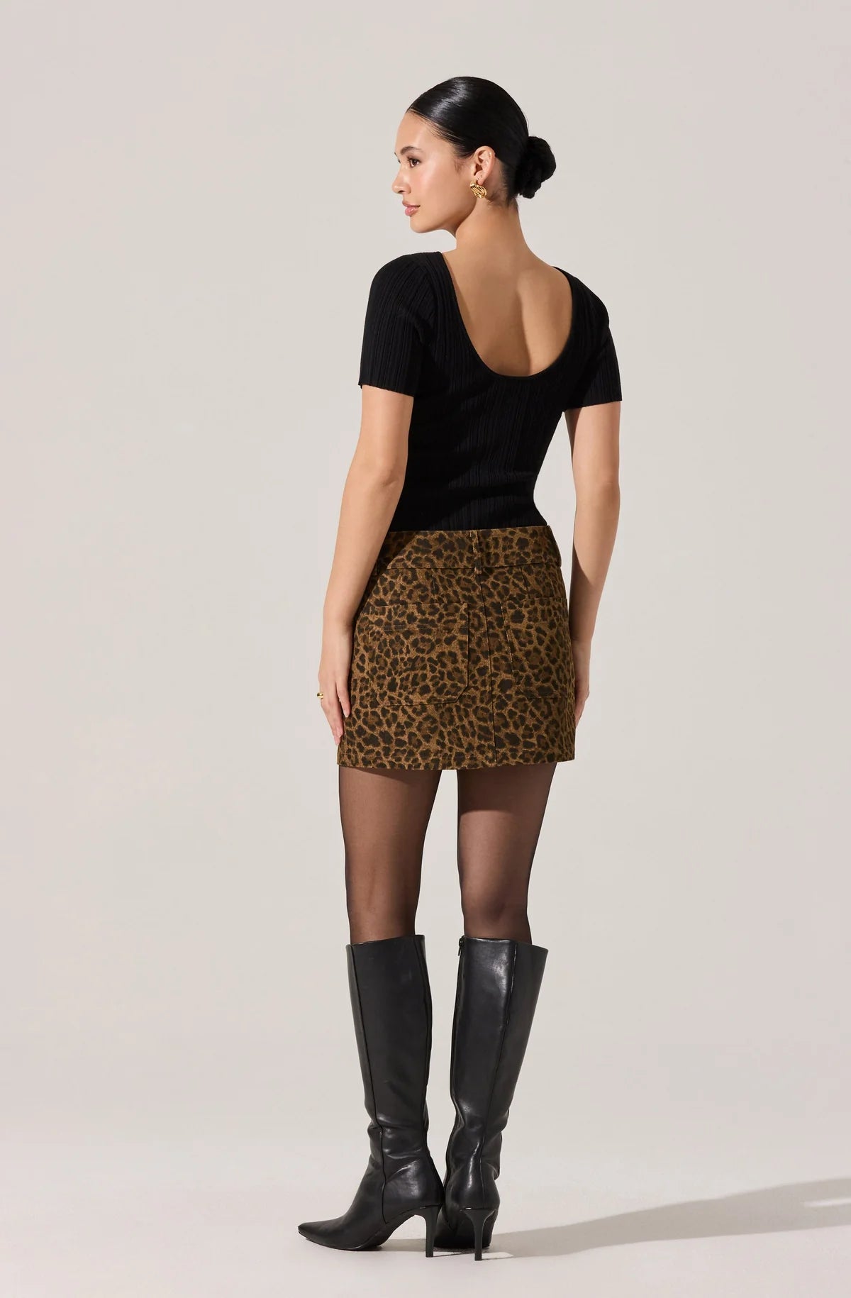 Woman wearing a black top and leopard print skirt with black boots on a plain background