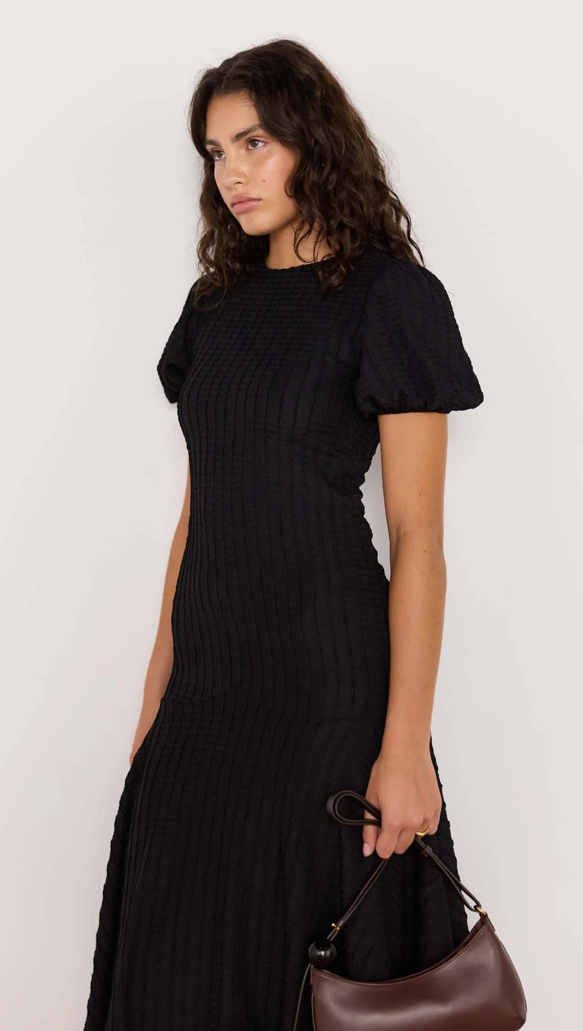 Woman wearing a black dress holding a brown handbag against a white background