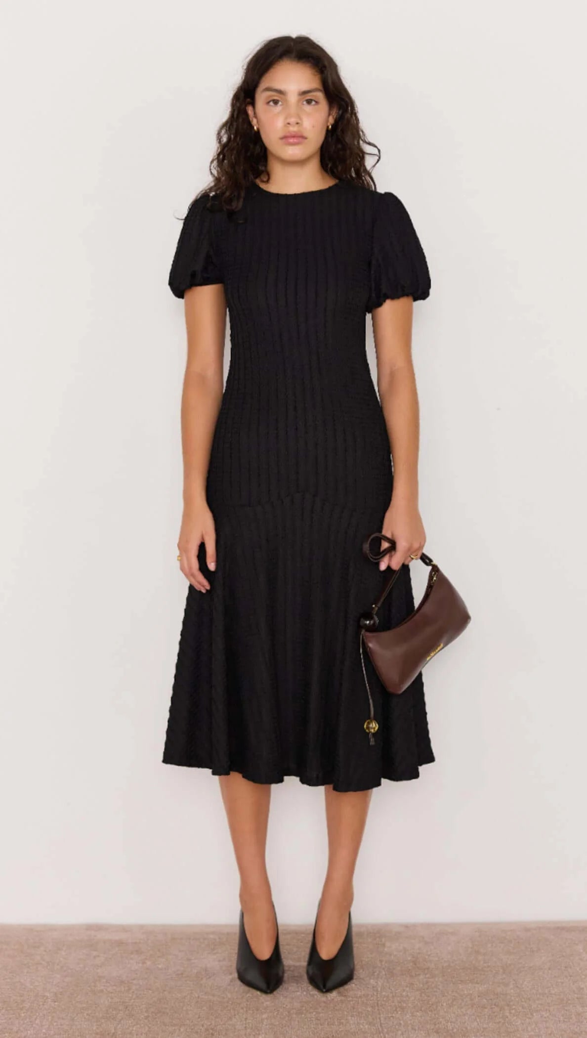 Woman wearing a black dress holding a brown handbag against a white background