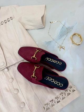 Red loafers with gold details on a light-colored coat with jewelry in the background