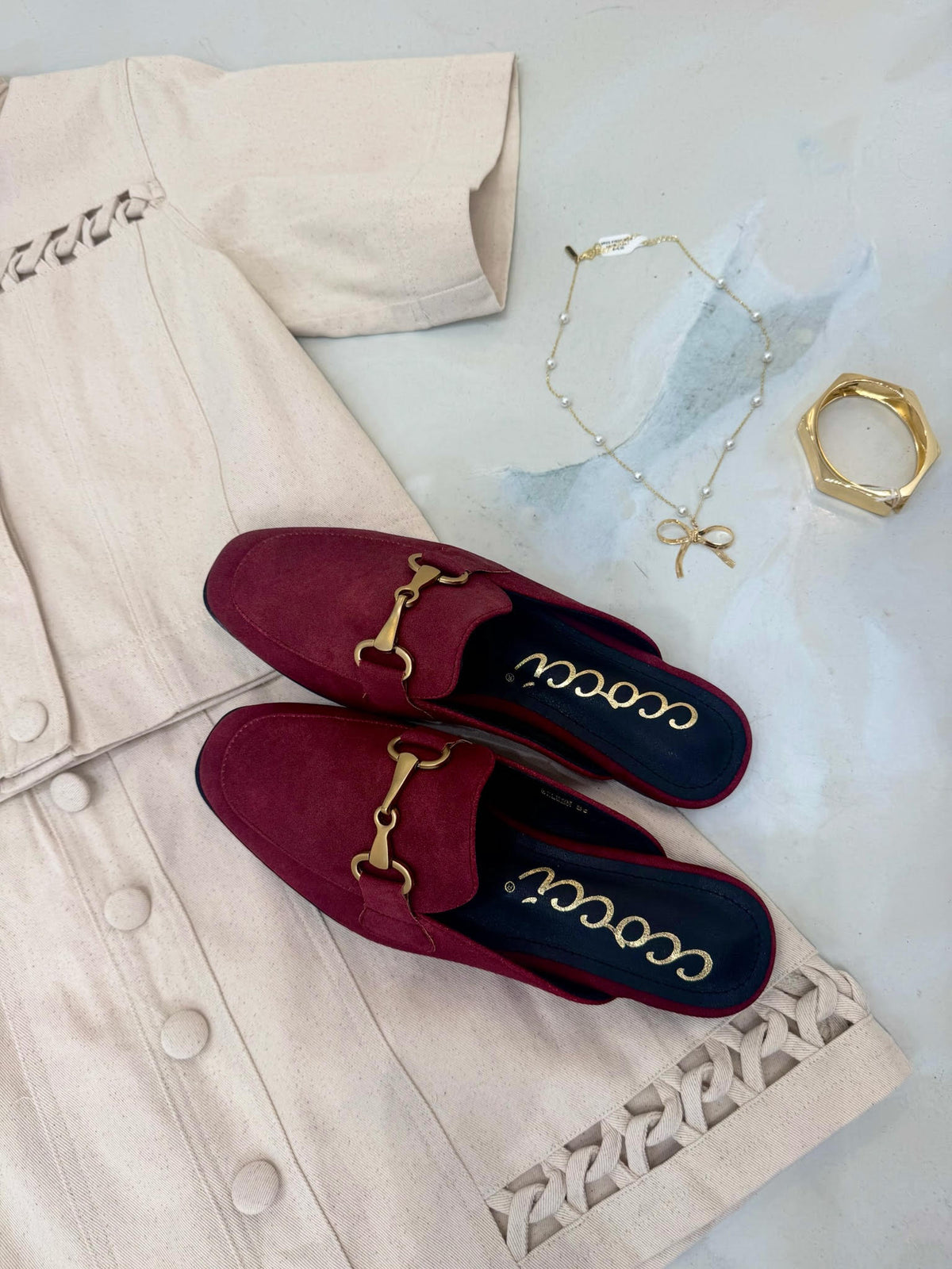 Red loafers with gold details on a light-colored coat with jewelry in the background