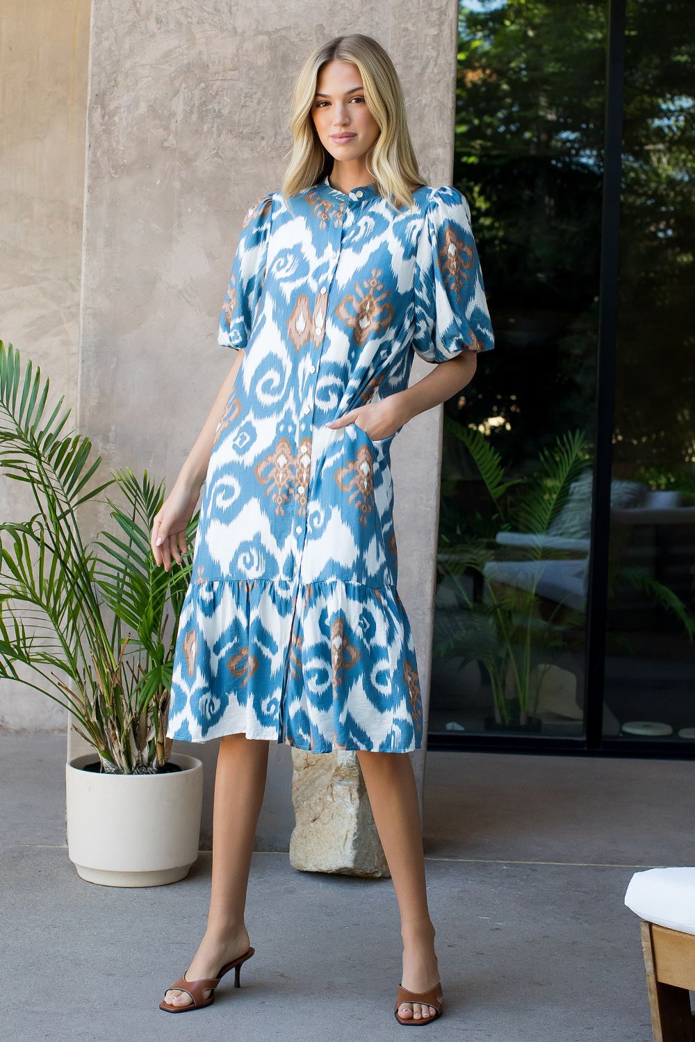 Woman wearing a blue patterned dress standing outdoors with plants and a building in the background.