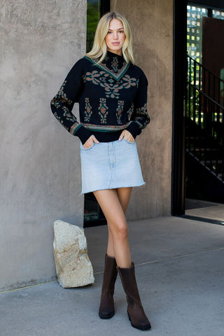 Woman wearing a dark sweater with a pattern, light blue denim skirt, and brown boots standing against a concrete wall.