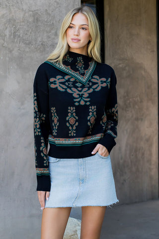 Woman wearing a black patterned sweater and light blue denim skirt against a gray wall.