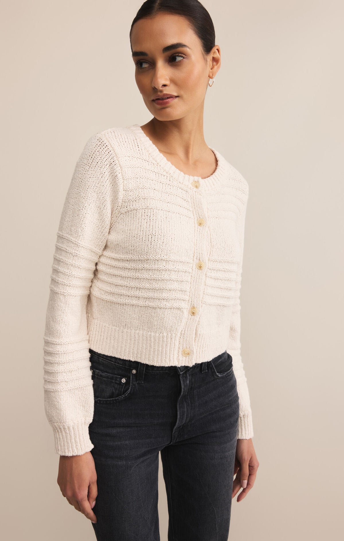 Woman wearing a cream knitted cardigan and dark jeans against a beige background