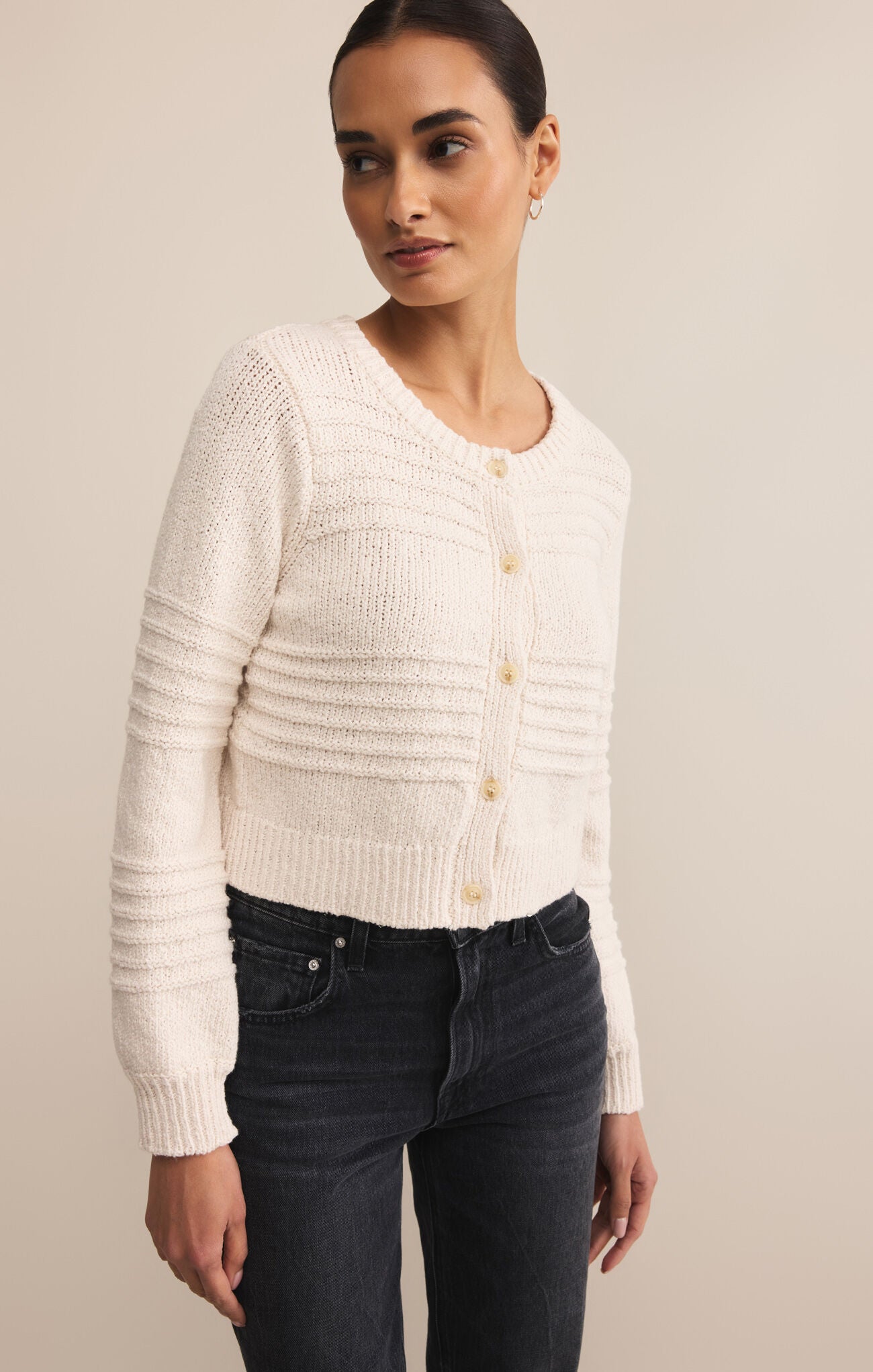 Woman wearing a cream knitted cardigan and dark jeans against a beige background